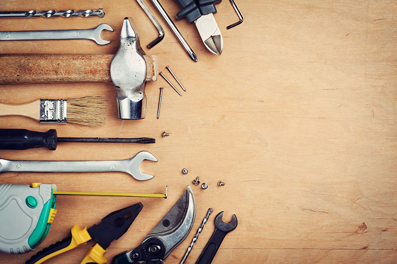 Assorted hand tools including wrenches, pliers, a hammer, a screwdriver, a paintbrush, a tape measure, and drill bits arranged on a wooden surface with empty space on the right.