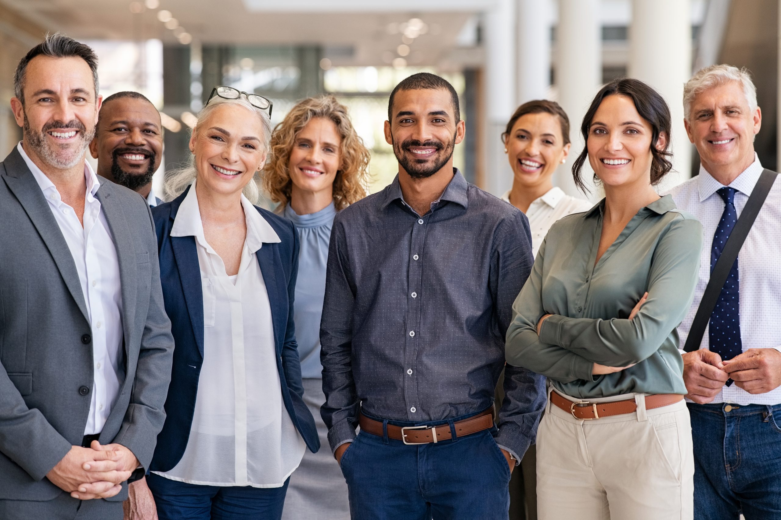 A diverse group of eight professionally dressed adults stand together indoors, smiling at the camera, suggesting a positive and collaborative work environment.